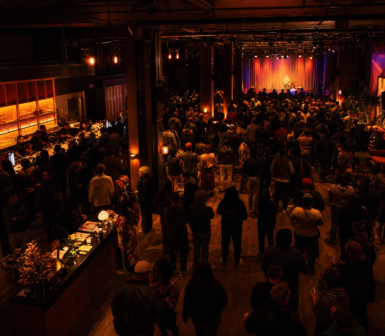 A wide view of a warm-lit cultural venue showing audience flow, stage attention, and room zones.